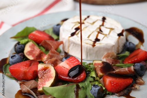 Pouring balsamic vinegar onto fresh salad with brie cheese, figs and berries on plate, closeup photo