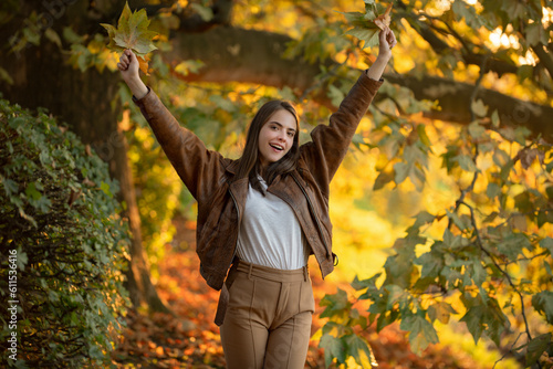 Happy woman holding autumn leafs on face in fall nature. Portrait of young woman with autumn maple leaves outdoor. Pretty girl play with fall leaf walking in park. Warm autumn. Fall season fashion.