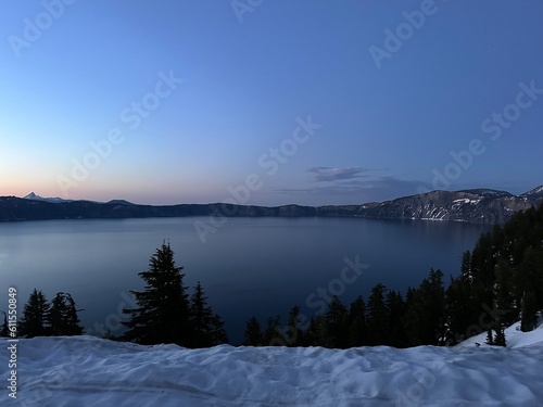 Crater Lake at twilight with fading light reflecting on the calm waters, silhouetted trees in foreground and distant volcanic crater rim in background.