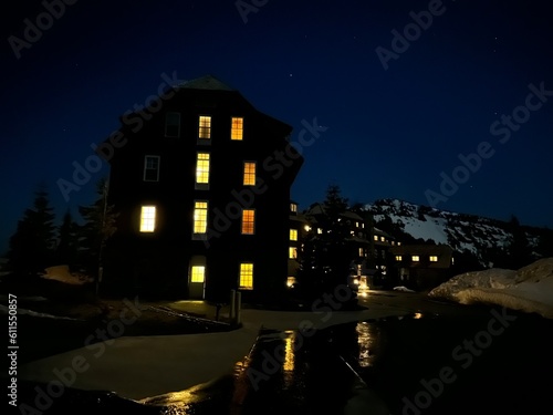 Crater Lake Lodge glowing at night with lit windows, surrounded by patches of snow, set under a deep blue star-filled sky in Oregon.