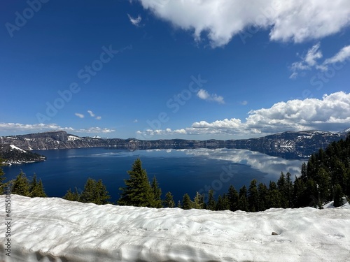 Crater Lake panorama from high elevation showing snow-covered ground, pine trees, and brilliant blue volcanic water under a partly cloudy sky