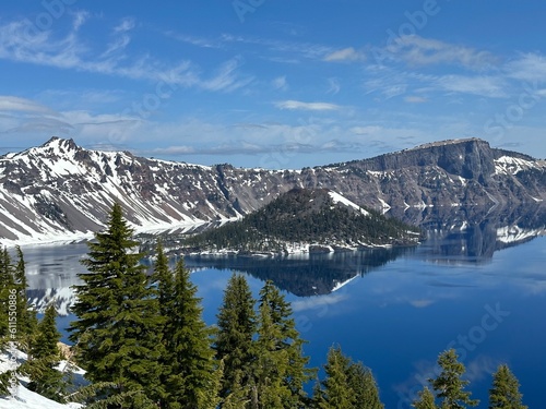 Bright sunny view of Crater Lake with Wizard Island surrounded by crystal-clear blue water, steep rocky crater walls, and patches of late-spring snow