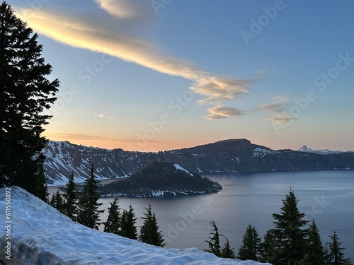 Sunset at Crater Lake with dramatic golden clouds reflecting over the calm blue volcanic lake surrounded by snow and forested ridges.