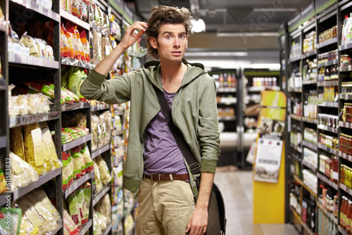 Confused, portrait and a man doing grocery shopping at a supermarket with doubt and thinking. Inflation, idea and a male shopper at a store with surprise at groceries, sale offer or customer deal photo