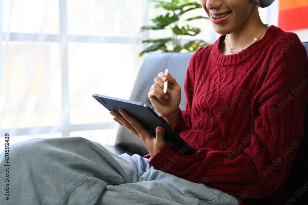 Happy young man in red sweater surfing internet on digital tablet, resting on couch in bright living room