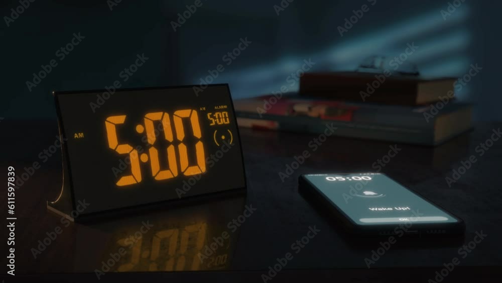Digital alarm clock with orange clockface and the smartphone waking up at 5 AM. The numbers on the clock screen changes from 4:59 no 5:00 AM. And the alarm goes off on the phone. Close up view.