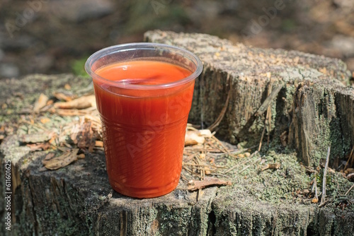 one plastic glass full of red tomato juice stands on a gray stump outdoors in nature