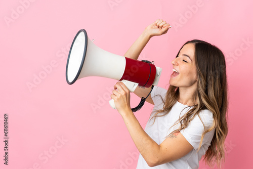 Young Romanian woman isolated on pink background shouting through a megaphone to announce something in lateral position