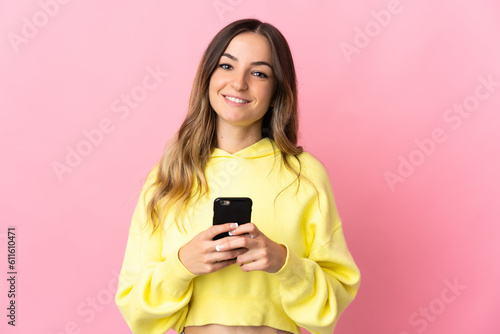 Young Romanian woman isolated on pink background looking at the camera and smiling while using the mobile