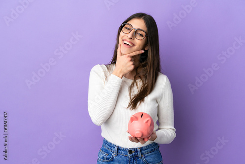 Young caucasian woman holding a piggybank isolated on purple background happy and smiling