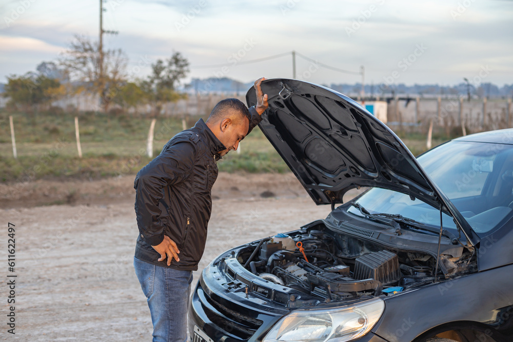 Young Latino man looking at the engine of his broken down car on the side of the road.