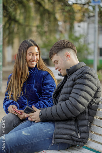 Couple of girl and boy sitting on the bench in a public park look at the mobile phone. © jroballo