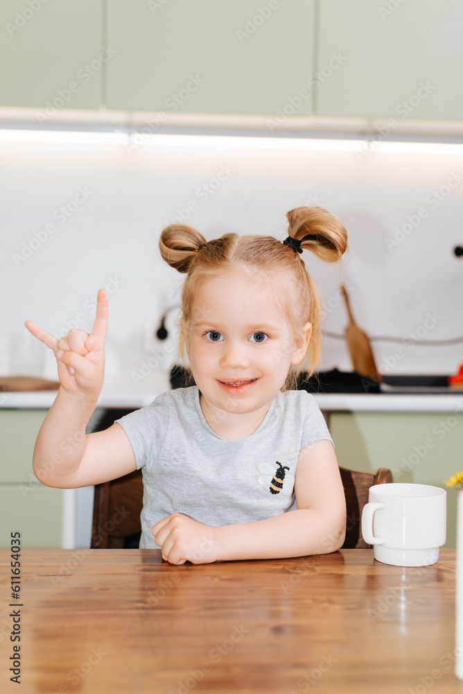 Funny child girl showing rock goat gesture while sitting at the table in the kitchen and looking at the camera, vertical angle Funny child girl showing rock goat gesture while sitting at the table in the kitchen and looking at the camera, vertical angle