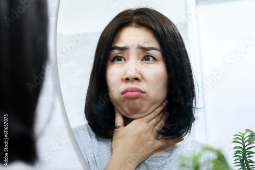 worried Asian woman checking her double chin under her lower jaw in front of a mirror, a sign of weight gain or obesity photo