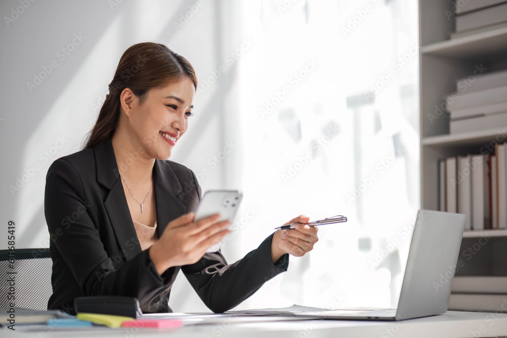 Attractive brunette Asian woman using mobile phone, smile and happy while sitting at working desk in modern office. Attractive brunette Asian woman using mobile phone, smile and happy while sitting at working desk in modern office.