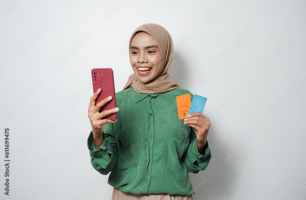 Smiling young Asian Muslim woman dressed in green shirt holding mobile phone and credit card, online shopping isolated over white background Smiling young Asian Muslim woman dressed in green shirt holding mobile phone and credit card, online shopping isolated over white background