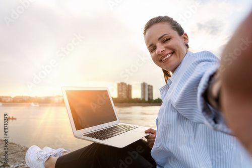 Smiling digital nomad woman working on laptop while sitting on waterfront taking selfie in front of cityscape and sunset.
