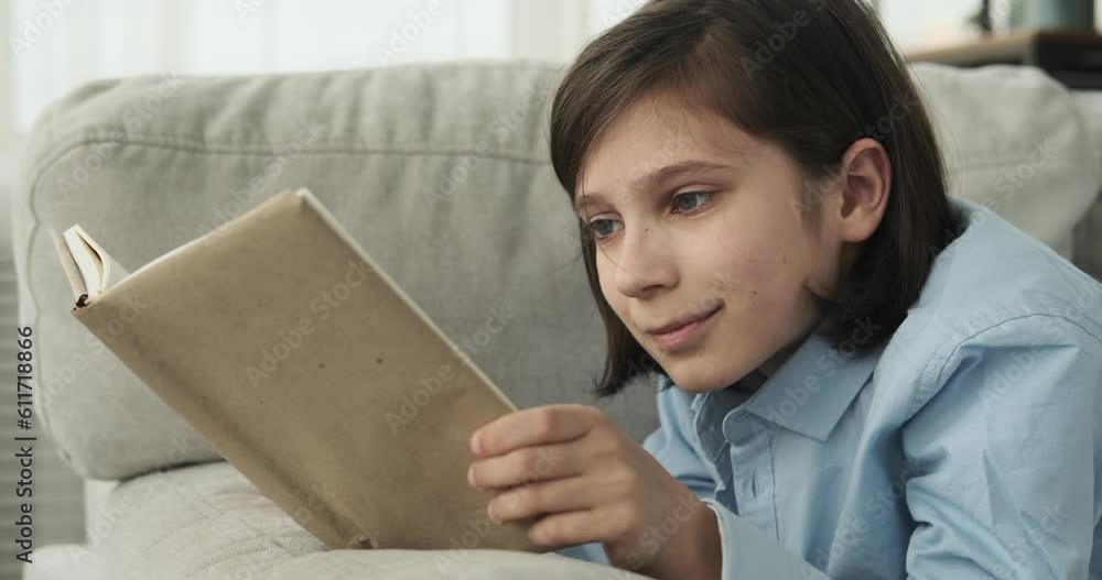 A boy as he finds solace in the enchanting world of books. The young boy is seen lying comfortably on the couch, a smile adorning his face, completely engrossed in the pages of a captivating book. A boy as he finds solace in the enchanting world of books. The young boy is seen lying comfortably on the couch, a smile adorning his face, completely engrossed in the pages of a captivating book.