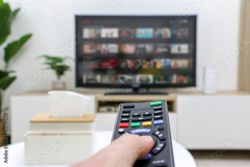 First person view of hand holding remote control, choosing what to watch on streaming platform displayed on television screen in background but out of focus, depicting home life