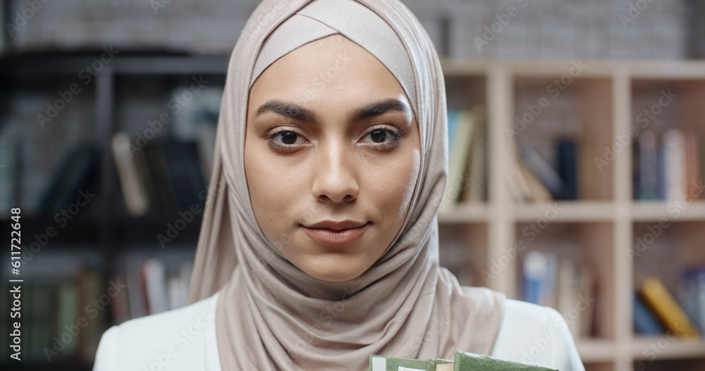 Female muslim student in library, holding books and looking at camera, girl wearing hijab spending time studying - modern Islam, student lifestyle concept close up Female muslim student in library, holding books and looking at camera, girl wearing hijab spending time studying - modern Islam, student lifestyle concept close up