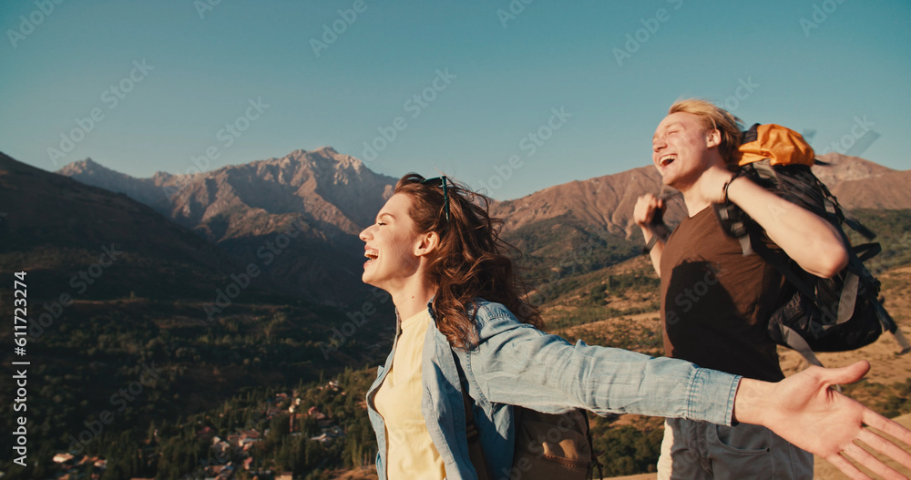 Friends hikers laughing with hands up, on a top of the mountain at sunset in mountains. Couple raising up hands on high rock in evening nature. Tourism, traveling and healthy lifestyle concept.