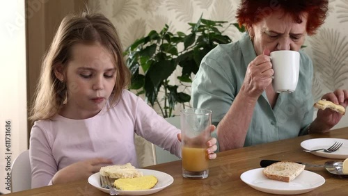 A little blonde girl is having breakfast in the kitchen with her beloved grandmother with an omelet, a sandwich with cheese and orange juice. Family traditions and friendship between generations. photo