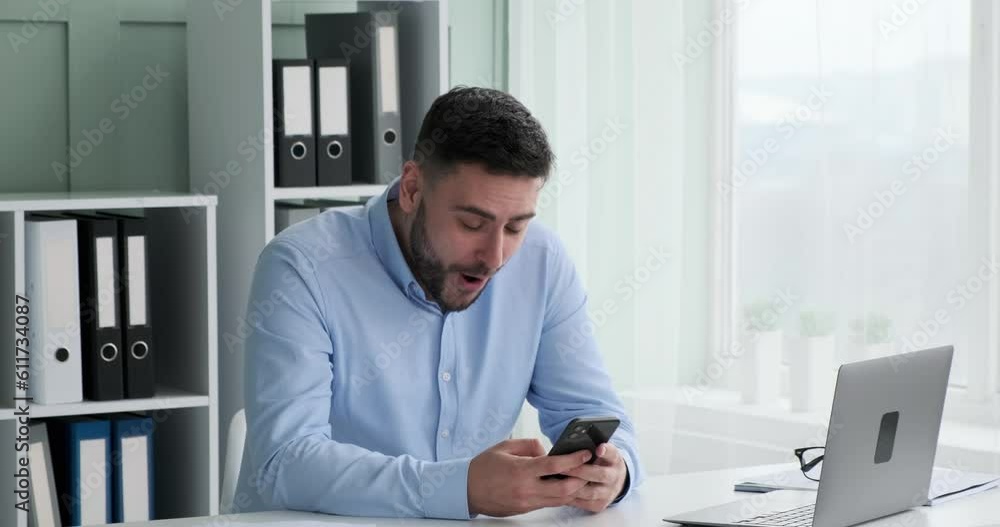 A Caucasian businessman in a shirt is sitting at his desk in the office, browsing his phone, and suddenly receives some good news. He smiles widely and starts typing excitedly on his phone. A Caucasian businessman in a shirt is sitting at his desk in the office, browsing his phone, and suddenly receives some good news. He smiles widely and starts typing excitedly on his phone.