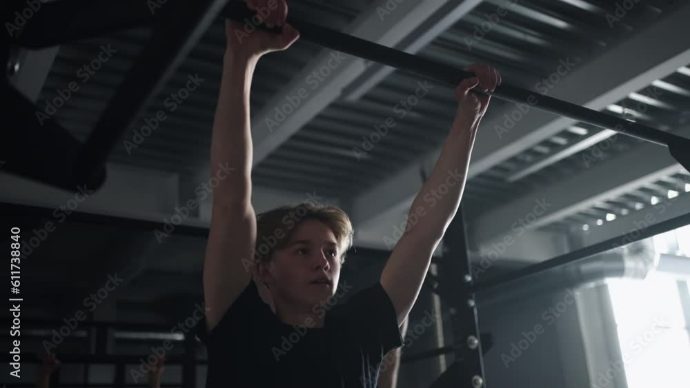 Young Athletic Guy Training In Gym, Doing Pull-Ups On Crossbar, Intense Group Workout Of Sportsmen Young Athletic Guy Training In Gym, Doing Pull-Ups On Crossbar, Intense Group Workout Of Sportsmen