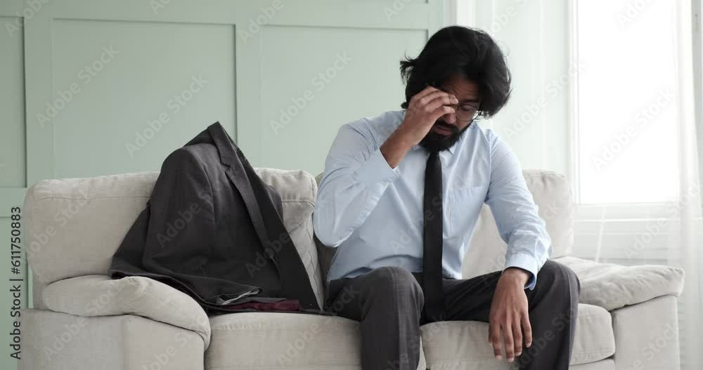 Indian businessman enters the frame, takes off his jacket and throws it on the couch, sits down, removes his glasses, and holds his head in distress, in a bad mood, feeling a headache. Indian businessman enters the frame, takes off his jacket and throws it on the couch, sits down, removes his glasses, and holds his head in distress, in a bad mood, feeling a headache.