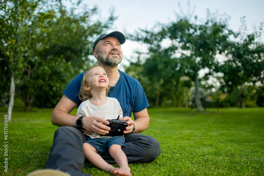 Father and son watching and navigating a drone. Cute toddler boy helping his father to operate a drone by remote control. Father and son watching and navigating a drone. Cute toddler boy helping his father to operate a drone by remote control.