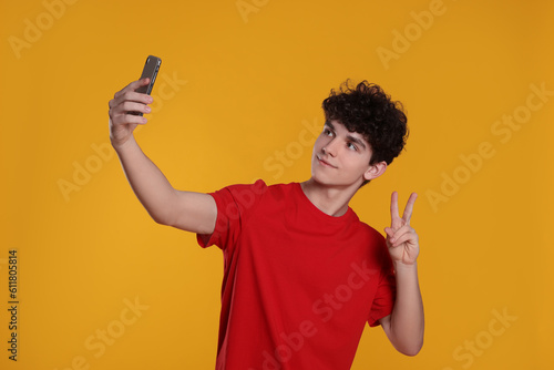 Teenage boy taking selfie and showing peace gesture on orange background