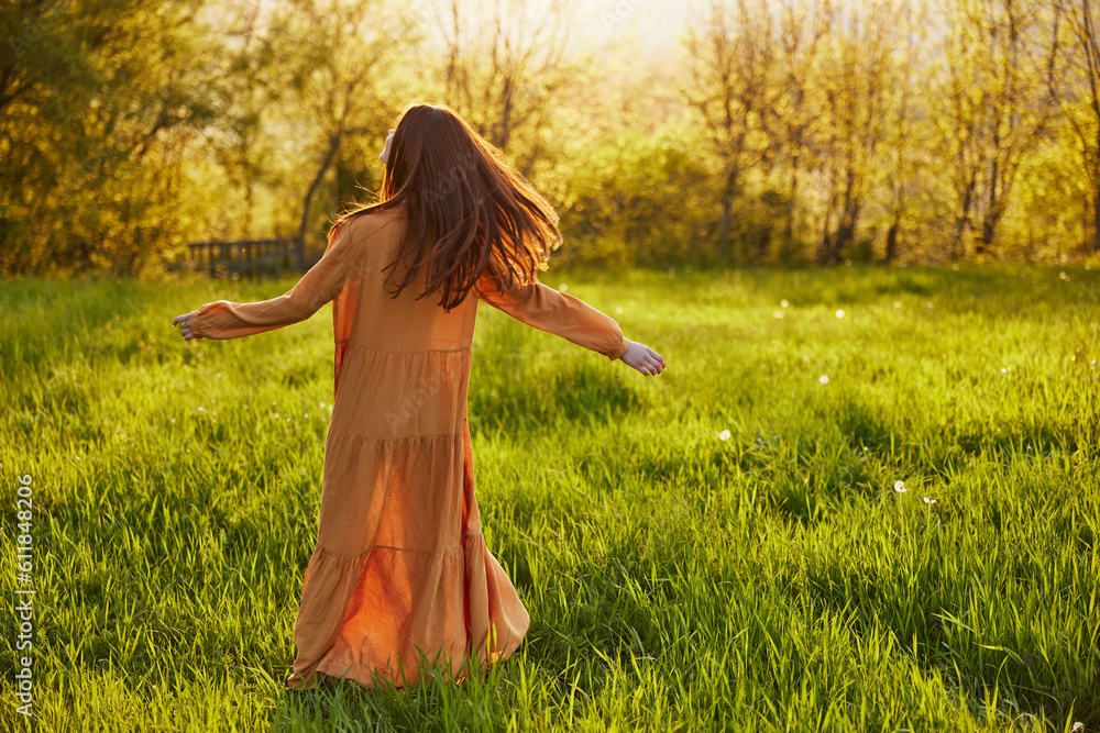 a relaxed slender woman enjoys the sunset standing in a green field with tall grass in an orange dress with her back to the camera, in warm summer weather. Horizontal photo a relaxed slender woman enjoys the sunset standing in a green field with tall grass in an orange dress with her back to the camera, in warm summer weather. Horizontal photo
