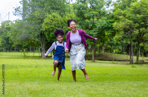 African American mother running barefoot on the grass lawn with her young daughter while having a summer picnic in the public park for wellbeing and happiness