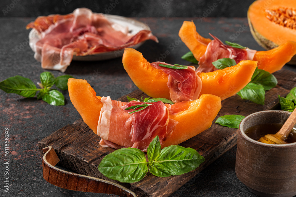 Prosciutto ham with melon cantaloupe slices, honey and basil on cutting board over dark background. Italian appetizer