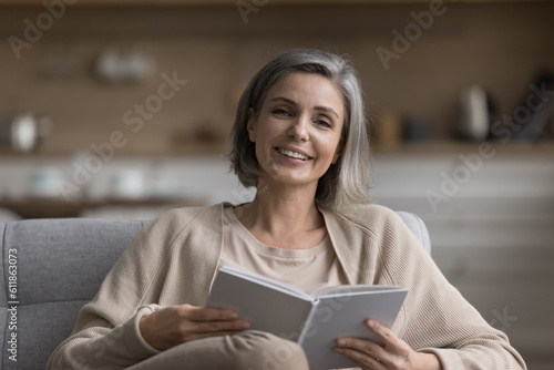 Happy grey haired mature paper book reader home hear shot portrait. Pretty middle aged woman holding textbook for studying new knowledge, education, looking at camera, smiling