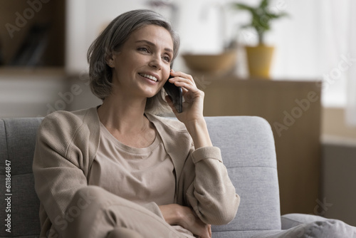 Happy positive mature woman talking on cellphone call, holding mobile phone at ear, relaxing on sofa, looking away, listening, smiling, laughing, enjoying communication, conversation, leisure