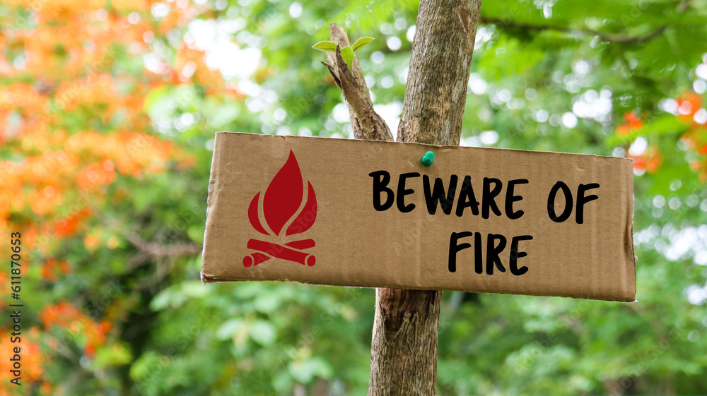 Brown paper label with drawing of bonfire and the text beware of fire on tree trunk with blurred background in camping site                              