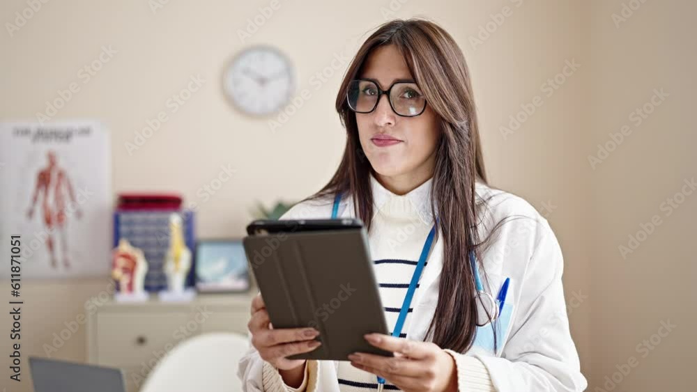Young beautiful hispanic woman doctor using touchpad saying no with head at clinic Young beautiful hispanic woman doctor using touchpad saying no with head at clinic