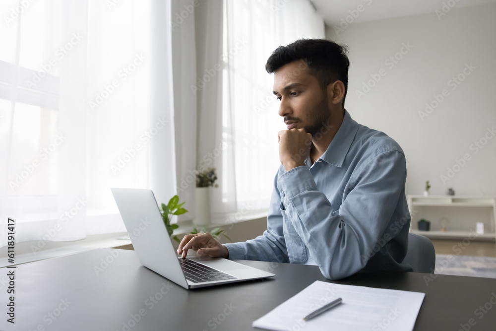 Serious busy young Indian freelance employee man typing on laptop, sitting at home office table, working at computer, using online application, Internet technology for job communication Serious busy young Indian freelance employee man typing on laptop, sitting at home office table, working at computer, using online application, Internet technology for job communication