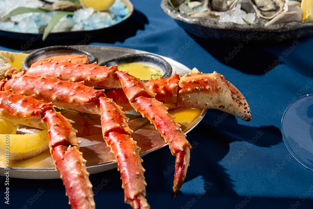 Closeup view of an elegant seafood assortment with prepared crab phalanges, lemon slices, sauces, and fresh oysters on a metal dish, presented on a blue tablecloth, single color blue backdrop. Closeup view of an elegant seafood assortment with prepared crab phalanges, lemon slices, sauces, and fresh oysters on a metal dish, presented on a blue tablecloth, single color blue backdrop.