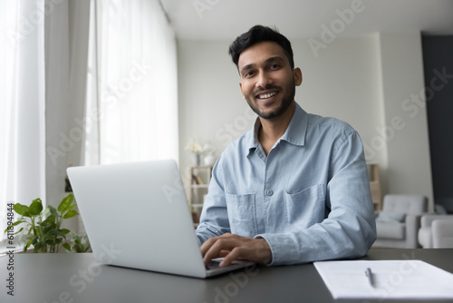 Happy young Indian freelance employee guy working at home, typing on laptop, looking at camera with toothy smile, enjoying internet communication. Remote business professional man portrait