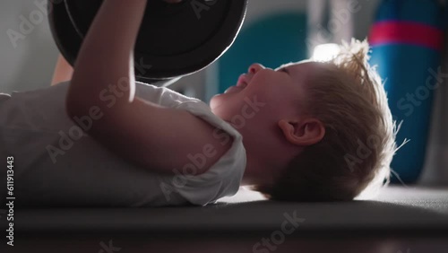 Motivated toddler tries hard to lift heavy barbell lying on floor in gym. Adorable little boy does difficult exercise with weight slow motion