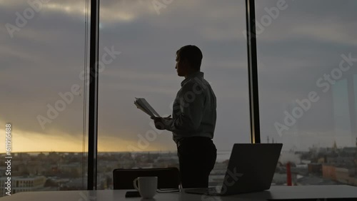 Businessman is standing at desk in office, holding important papers, looking out at beautiful sunset photo