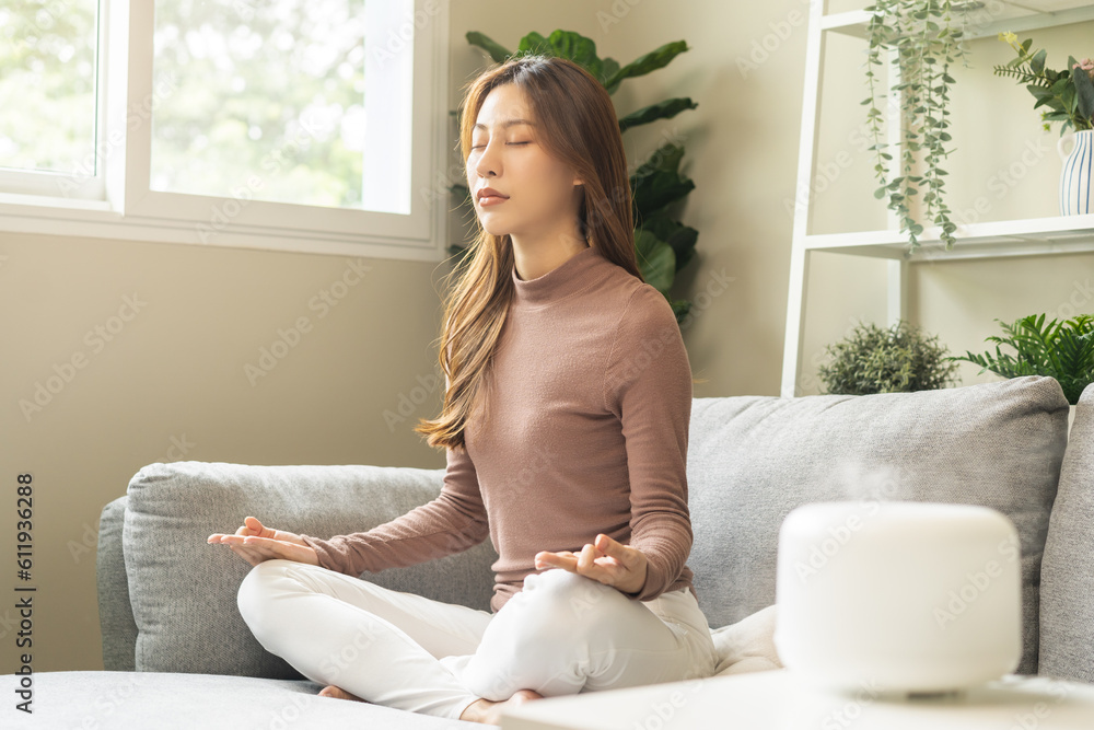 Air humidifier, calm asian young woman sitting on couch lotus pose put hands practice meditation do yoga exercise at home. Aromatherapy steam scent from essential oil diffuser in living room at home. Air humidifier, calm asian young woman sitting on couch lotus pose put hands practice meditation do yoga exercise at home. Aromatherapy steam scent from essential oil diffuser in living room at home.