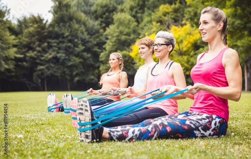 Women sitting on green grass in a row exercising with blue elastic band at park