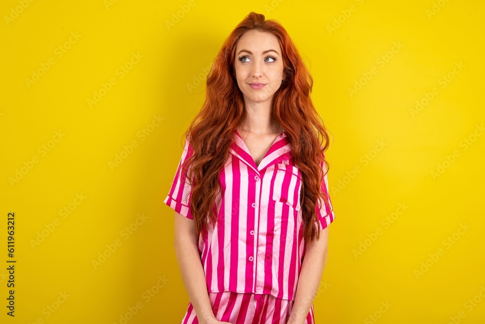 Young red haired woman wearing pink pyjama over yellow studio background looking aside into empty space thoughtful Young red haired woman wearing pink pyjama over yellow studio background looking aside into empty space thoughtful