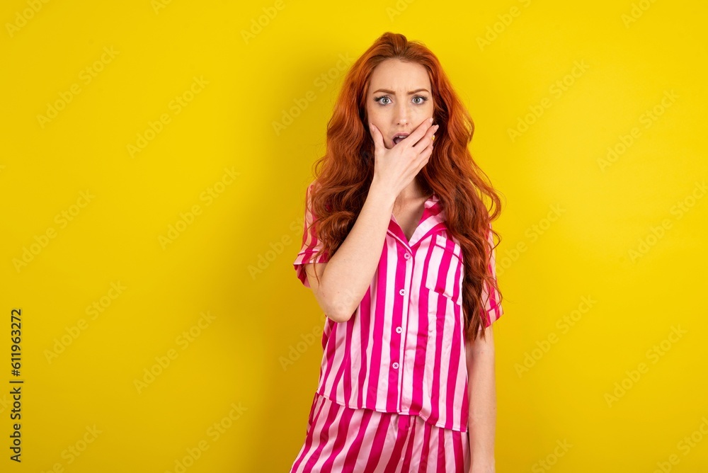 Emotional Young red haired woman wearing pink pyjama over yellow studio background gasps from astonishment, covers opened mouth with palm, looks shocked at camera. Emotional Young red haired woman wearing pink pyjama over yellow studio background gasps from astonishment, covers opened mouth with palm, looks shocked at camera.