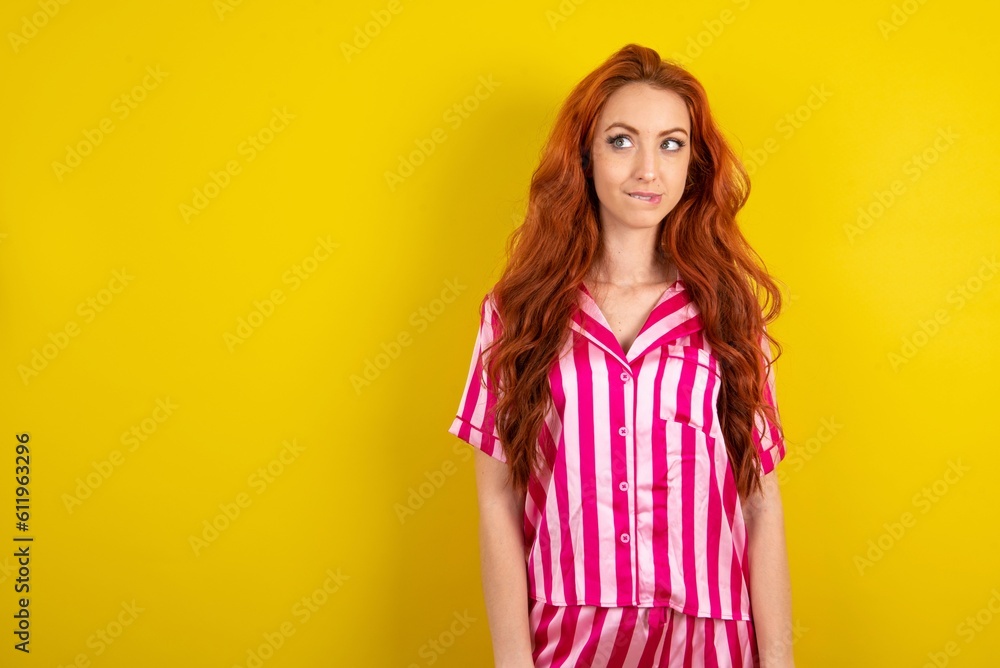 Amazed Young red haired woman wearing pink pyjama over yellow studio background bitting lip and looking tricky to empty space. Amazed Young red haired woman wearing pink pyjama over yellow studio background bitting lip and looking tricky to empty space.