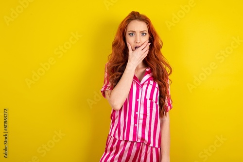 Emotional Young red haired woman wearing pink pyjama over yellow studio background gasps from astonishment, covers opened mouth with palm, looks shocked at camera.