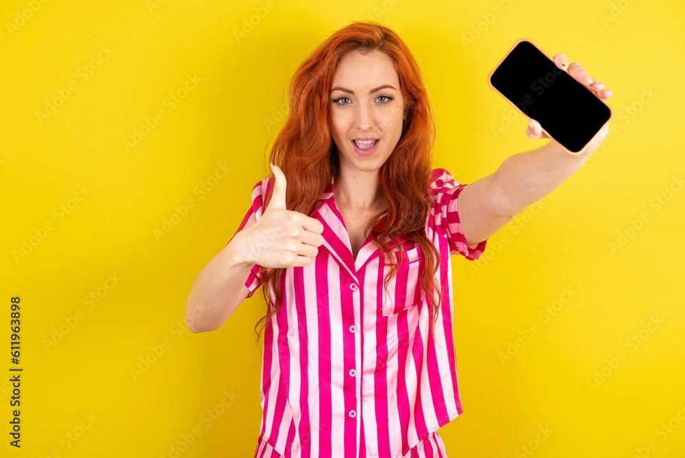 Portrait Young red haired woman wearing pink pyjama over yellow studio background holding in hands cell showing giving black screen thumb up Portrait Young red haired woman wearing pink pyjama over yellow studio background holding in hands cell showing giving black screen thumb up